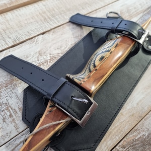 May include: A wooden bow with a leather grip and black leather straps. The bow is secured within a black leather sheath, with a silver buckle fastening the strap. The wood has a natural grain pattern.