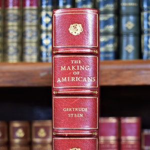 May include: A red leather-bound book titled "The Making of Americans" by Gertrude Stein. The book has gold accents and the text "Paris 1925" on the bottom.