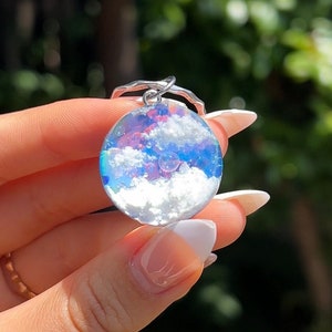 Puede incluir: Un charm redondo de plata con una cúpula de resina transparente que contiene un cielo azul y blanco con nubes. El charm está unido a un anillo de plata.