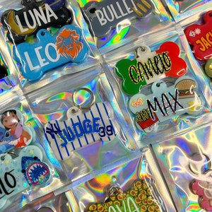 Op de afbeelding: Een verzameling kleurrijke hondenpenningen met verschillende namen en ontwerpen. De penningen zijn gemaakt van plastic en hebben de vorm van een bot. Sommige penningen hebben glitter, andere hebben cartoonfiguren en sommige hebben simpele tekst. De penningen zijn verpakt in doorzichtige plastic zakken.