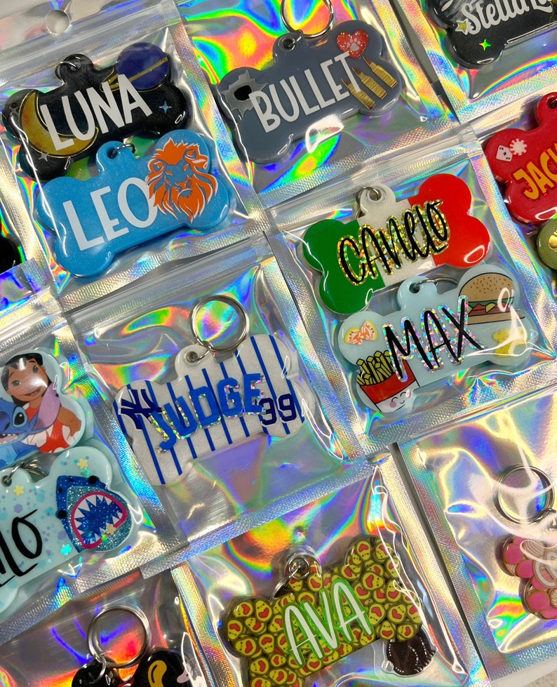 Op de afbeelding: Een verzameling kleurrijke hondenpenningen met verschillende namen en ontwerpen. De penningen zijn gemaakt van plastic en hebben de vorm van een bot. Sommige penningen hebben glitter, andere hebben cartoonfiguren en sommige hebben simpele tekst. De penningen zijn verpakt in doorzichtige plastic zakken.