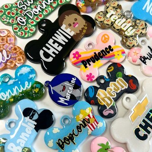 Op de afbeelding: Een assortiment kleurrijke hondenpenning-charms met verschillende ontwerpen en namen. De charms zijn gemaakt van hars en hebben een verscheidenheid aan thema's, waaronder cartoonfiguren, eten en natuur. De charms zijn allemaal in de vorm van een bot of een cirkel.