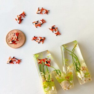 May include: A resin letter M with a clear background, filled with green and brown plants, white sand, and small orange and black fish. The letter is sitting on a white surface with a US penny and several small orange and black fish.