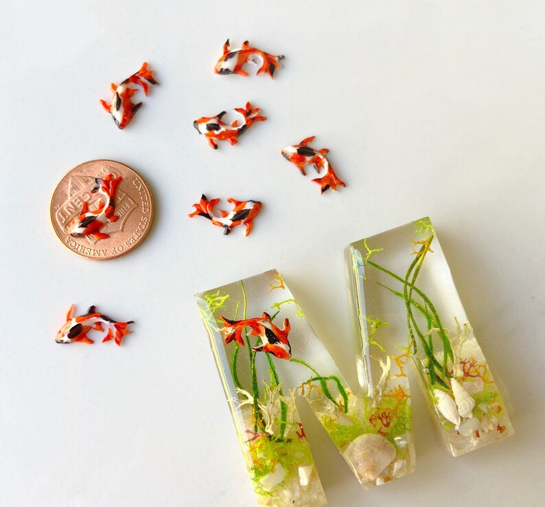 May include: A resin letter M with a clear background, filled with green and brown plants, white sand, and small orange and black fish. The letter is sitting on a white surface with a US penny and several small orange and black fish.