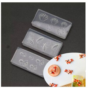 May include: Three clear silicone molds for making resin crafts. The molds feature different designs: seashells, flowers, and goldfish. The molds are shown next to a plate with several small, orange and black resin goldfish.