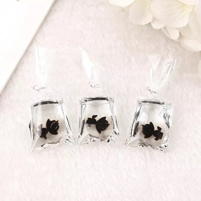 May include: Three clear plastic bags with black butterfly charms inside. The bags are hanging from a white surface.