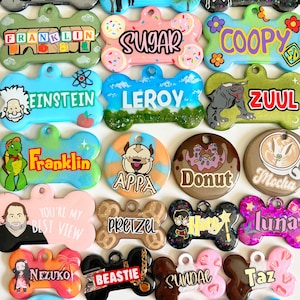 Op de afbeelding: Een verzameling van 20 hondenpenningen met verschillende ontwerpen en namen. De penningen zijn botvormig en gemaakt van hars met glitteraccenten. Sommige van de namen zijn Opie, Franklin, Leroy, Zuul, Apppa, Donut, Mocha, Pretzel, Luna, Nezuko, Beastie, Sundae, Taz, Cannoli, Sweetie Pie, Tater Tot, Moco en Stella.