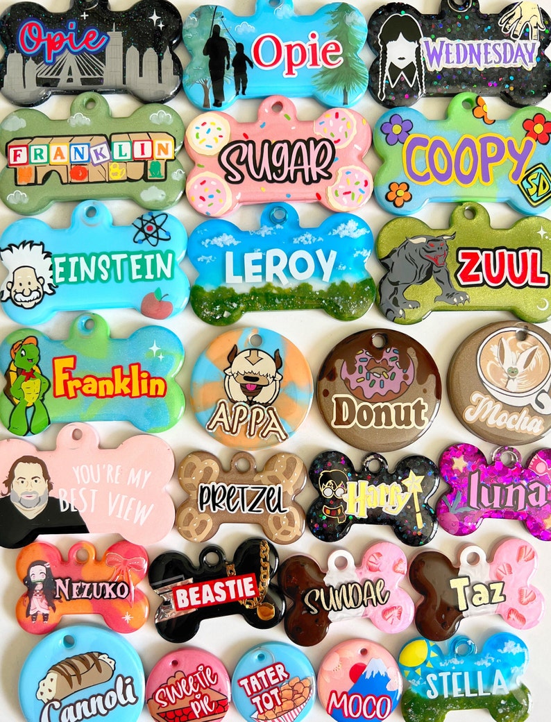 Op de afbeelding: Een verzameling van 20 hondenpenningen met verschillende ontwerpen en namen. De penningen zijn botvormig en gemaakt van hars met glitteraccenten. Sommige van de namen zijn Opie, Franklin, Leroy, Zuul, Apppa, Donut, Mocha, Pretzel, Luna, Nezuko, Beastie, Sundae, Taz, Cannoli, Sweetie Pie, Tater Tot, Moco en Stella.