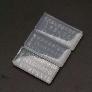 Puede incluir: Tres moldes de silicona transparentes para hacer manualidades de resina. Los moldes son rectangulares y presentan letras y números en relieve. El primer molde tiene los números del 1 al 0, el segundo molde tiene las letras de la a a la z, y el tercer molde tiene las letras de la a a la z en mayúsculas.