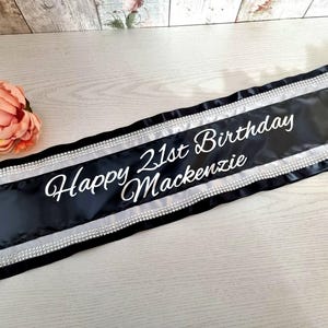 Könnte beinhalten: Schwarzes Satin-Geburtstagsschärpe mit silbernen Akzenten und dem Text "Happy 21st Birthday Mackenzie" in weißer Schrift. Das Schärpe ist mit einem silbernen Rand und Strasssteinen verziert. Eine pfirsichfarbene Blume ist im Hintergrund.