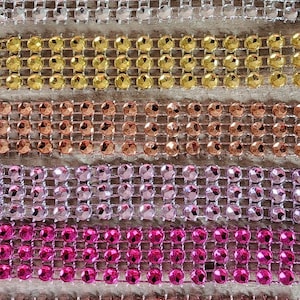 May include: Six horizontal strips of rhinestone trim arranged in a vertical stack. Each strip features three rows of round, faceted crystals set in a metallic mesh. The colors include silver, gold, copper, light pink, hot pink, and bright red.