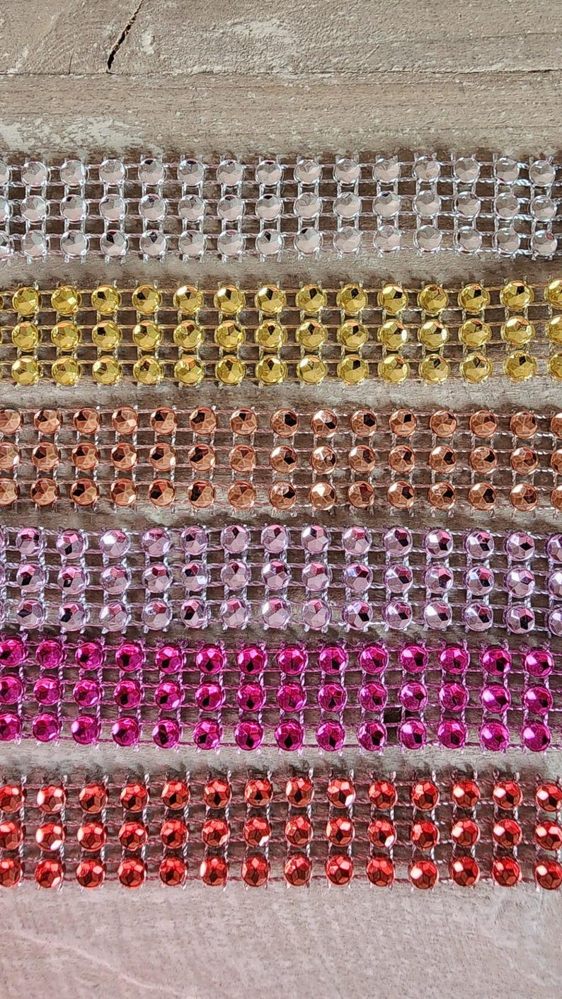 May include: Six horizontal strips of rhinestone trim arranged in a vertical stack. Each strip features three rows of round, faceted crystals set in a metallic mesh. The colors include silver, gold, copper, light pink, hot pink, and bright red.