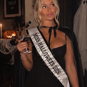 May include: A black mini dress with a matching black cape and a white satin sash featuring the black text MISS HALLOWEEN 2026. The sash is worn diagonally across the torso, and the cape is secured at the neck with a black ribbon tie.