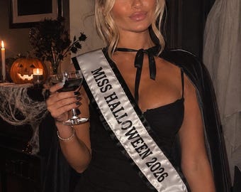 Puede incluir: Un vestido corto negro con una capa negra a juego y una banda de satén blanco con el texto negro MISS HALLOWEEN 2026. La banda se lleva en diagonal sobre el torso y la capa se sujeta al cuello con una cinta negra.