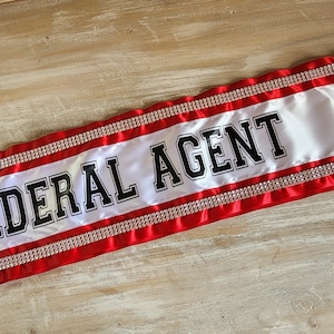 May include: A white satin sash featuring the text FEDERAL AGENT in bold black lettering. The sash is bordered by red satin fabric strips and two rows of sparkling silver rhinestones running horizontally along the top and bottom edges.
