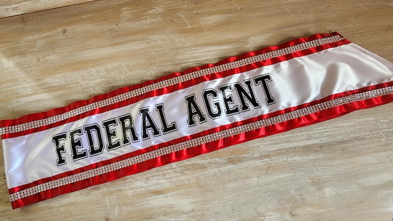 May include: A white satin sash featuring the text FEDERAL AGENT in bold black lettering. The sash is bordered by red satin fabric strips and two rows of sparkling silver rhinestones running horizontally along the top and bottom edges.