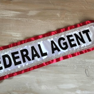May include: A white satin sash featuring the text FEDERAL AGENT in bold black capital letters. The sash is bordered by red satin fabric and accented with a row of clear, sparkling rhinestones along both edges of the central white panel.