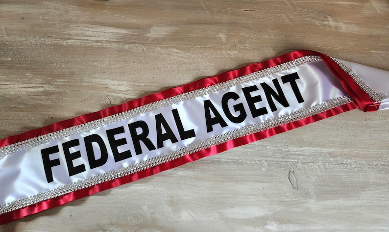 May include: A white satin sash featuring the text FEDERAL AGENT in bold black capital letters. The sash is bordered by red satin fabric and accented with a row of clear, sparkling rhinestones along both edges of the central white panel.