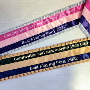 May include: Two satin sashes with gold and white lettering. The top sash is silver, pink, and purple. The bottom sash is gold, black, and navy blue. Both sashes say "Cambridge and Newmarket Polo Club" and "Best Playing Pony 2022".