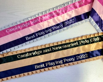 Puede incluir: Dos bandas de premio de satén con bordes plisados. La banda superior es rosa, rosa claro y azul oscuro con texto blanco Cambridge and Newmarket Polo Club Best Playing Pony 2022. La banda inferior es verde oscuro, beige y azul oscuro con texto dorado.