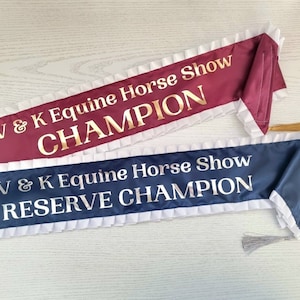 May include: Two satin horse show ribbons with white ruffled trim. The top ribbon is burgundy with gold lettering that reads "V & K Equine Horse Show CHAMPION". The bottom ribbon is navy blue with silver lettering that reads "V & K Equine Horse Show RESERVE CHAMPION".