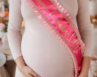Puede incluir: Una banda de satén rosa brillante con texto dorado en cursiva que dice Mumma to baby Lillie. La banda está adornada con bordes de tela rosa claro con volantes y una fina línea de brillantes diamantes de imitación a ambos lados del texto.