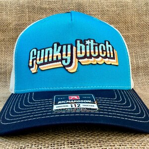 May include: A blue and black trucker hat with a white mesh back. The front of the hat features a retro-style embroidered patch that says "Funky Bitch". The hat is adjustable and has a Richardson 112 label.