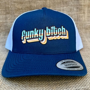 May include: A navy blue and white trucker hat with a retro-style "Funky Bitch" embroidered on the front. The hat has a white mesh back and a snap closure.