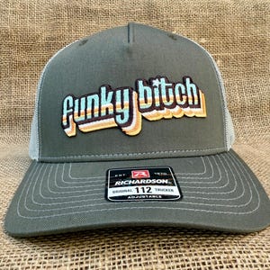 May include: A green and white trucker hat with a retro-style embroidered patch that says "Funky Bitch". The hat is adjustable and has a Richardson 112 label.