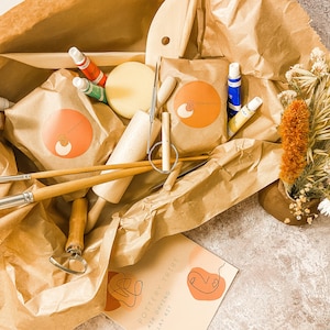 May include: A pottery kit with various tools, paints, and clay, arranged in a brown paper package. Includes brushes, a rolling pin, and a kit labeled "Pottery Tribe Air Drying Clay Kit". A small vase with dried flowers is also present.