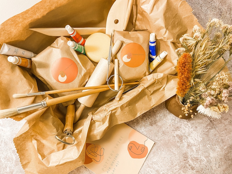 May include: A pottery kit with various tools, paints, and clay, arranged in a brown paper package. Includes brushes, a rolling pin, and a kit labeled "Pottery Tribe Air Drying Clay Kit". A small vase with dried flowers is also present.