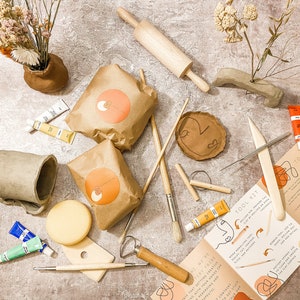 May include: A collection of pottery tools and supplies, including paint tubes, brushes, a rolling pin, and clay. The image also features a handmade mug, vases, and packaged clay. A brochure with the text "TOOL KIT" is also visible.