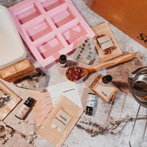May include: A selection of soap-making ingredients and tools, including a pink silicone mould, essential oil bottles, dried flowers, and instructions. The image features a wooden spoon filled with rose petals, soap bars, and a container labelled "Soak."