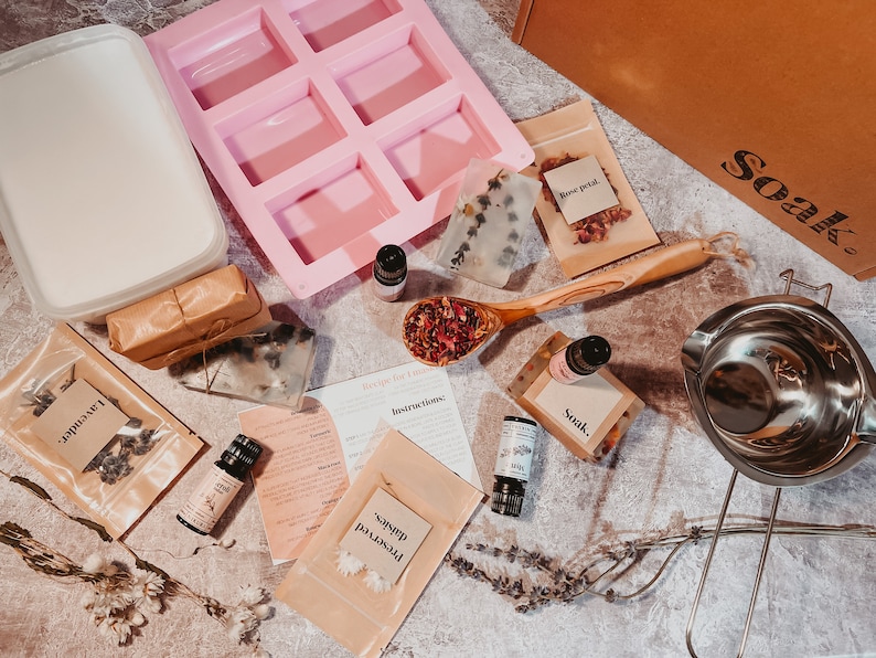 May include: A flat lay of soap-making supplies, including a pink silicone mold, essential oil bottles, dried flowers, and instructions. The image also features a container of soap base, packaged soap bars, and a wooden spoon filled with rose petals. The word "Soak." is visible.
