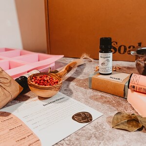 May include: A collection of natural skincare products, including a pink soap mold, essential oil bottles, a wooden scoop filled with red berries, and a bar of soap. The image also shows a recipe card and a brown box.