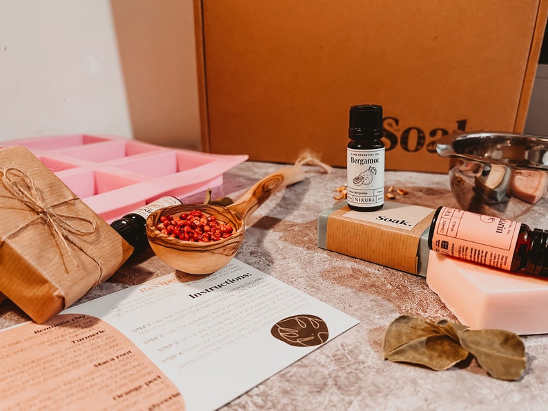 May include: A collection of natural skincare products, including a pink soap mold, essential oil bottles, a wooden scoop filled with red berries, and a bar of soap. The image also shows a recipe card and a brown box.