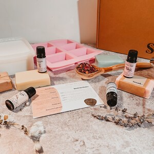 May include: A soap-making kit with various components, including essential oils, a pink silicone mold, soap bars, and a wooden spoon filled with dried flowers. The kit also includes a container, instructions, and a gift-wrapped package.