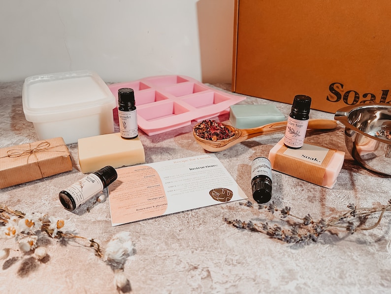 May include: A soap-making kit with various components, including essential oils, a pink silicone mold, soap bars, and a wooden spoon filled with dried flowers. The kit also includes a container, instructions, and a gift-wrapped package.