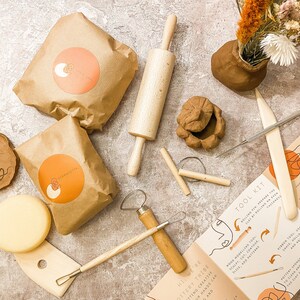 May include: A pottery kit with clay, tools, and instructions. The kit includes clay in brown paper bags with orange labels, a wooden rolling pin, and various sculpting tools. A small clay pot with dried flowers is also present.
