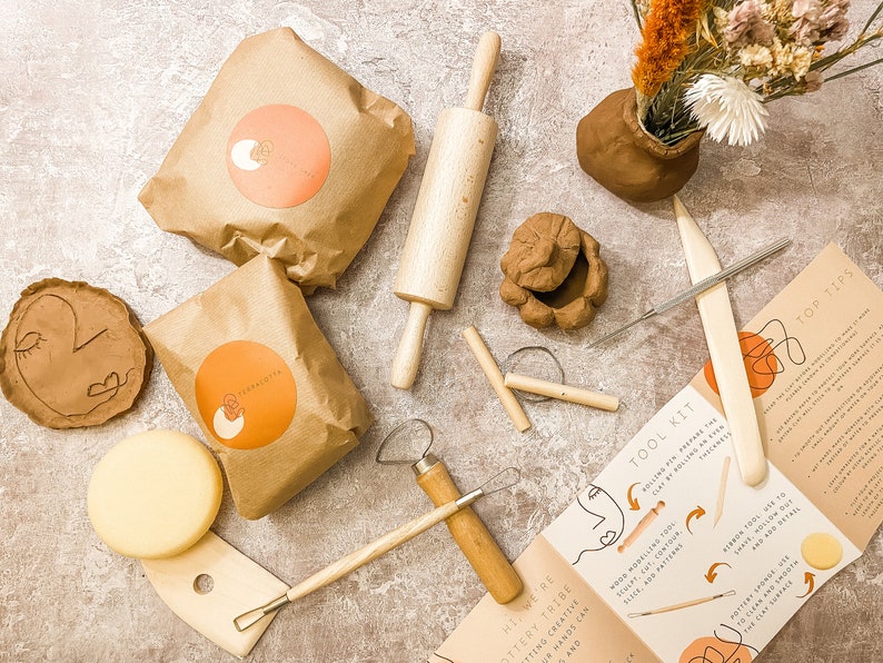 May include: A pottery kit with clay, tools, and instructions. The kit includes clay in brown paper bags with orange labels, a wooden rolling pin, and various sculpting tools. A small clay pot with dried flowers is also present.
