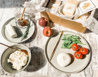 May include: A cheese-making kit with ingredients and recipe card, alongside plates of fresh ingredients like tomatoes, basil, and peaches. Includes a jar of honey and a sprig of rosemary. The kit is in a cardboard box.