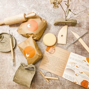May include: A pottery tool kit with various items, including a wooden rolling pin, clay, a small vase, and several wooden tools. The kit also includes a guide with instructions and illustrations. The items are arranged on a textured surface.