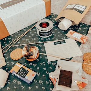 May include: A s'mores kit laid out with a box, toasting tin, biscuits, marshmallows, chocolate, and instructions. The box is white with a brown lid and a tree pattern. A toasted marshmallow sits on a biscuit. Measurements are in cm and grams.