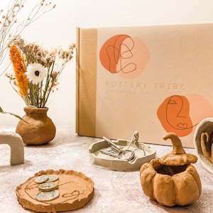 May include: A Pottery Tribe air-drying clay kit with handmade clay objects. The objects include a vase with dried flowers, a pumpkin-shaped container, a tray with coins, a small dish with keys, and a decorative sculpture.
