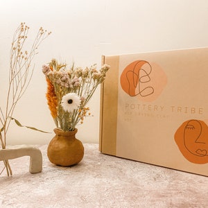 May include: A craft kit box labeled "Pottery Tribe Air Drying Clay Kit" with abstract face illustrations. Beside it are dried flowers in a small brown vase and a clay sculpture. The background is a neutral color.