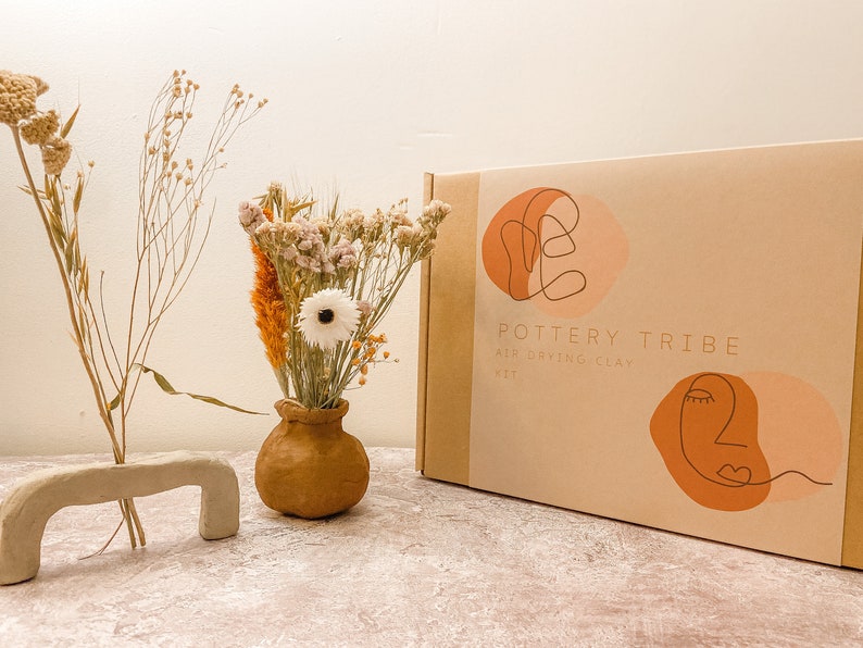 May include: A craft kit box labeled "Pottery Tribe Air Drying Clay Kit" with abstract face illustrations. Beside it are dried flowers in a small brown vase and a clay sculpture. The background is a neutral color.