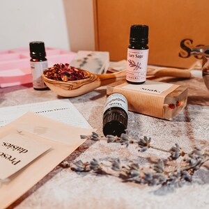 May include: A collection of essential oil bottles, a wooden spoon filled with dried flowers, and a bar of soap. The scene includes a pink soap mold, instructions, and a sprig of lavender, suggesting a DIY spa kit.