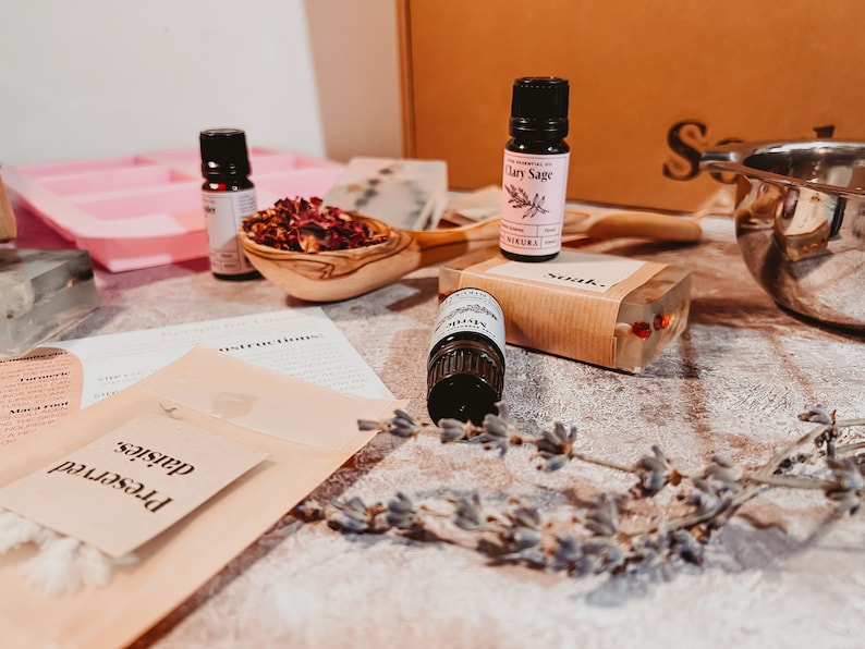 May include: A collection of essential oil bottles, a wooden spoon filled with dried flowers, and a bar of soap. The scene includes a pink soap mold, instructions, and a sprig of lavender, suggesting a DIY spa kit.