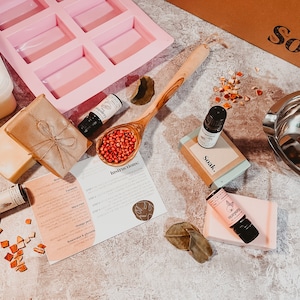 May include: A selection of soap-making materials arranged on a surface. The image features a pink silicone mould, essential oil bottles, a wooden spoon with red peppercorns, soap bars, and a stainless steel double boiler. The word "Soak" is printed on a cardboard box.
