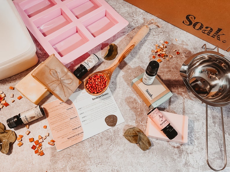 May include: A flat lay of soap-making supplies. Includes a pink silicone mold, essential oil bottles, a wooden spoon filled with red peppercorns, bars of soap, and a stainless steel double boiler. The word "Soak" is printed on a cardboard box.